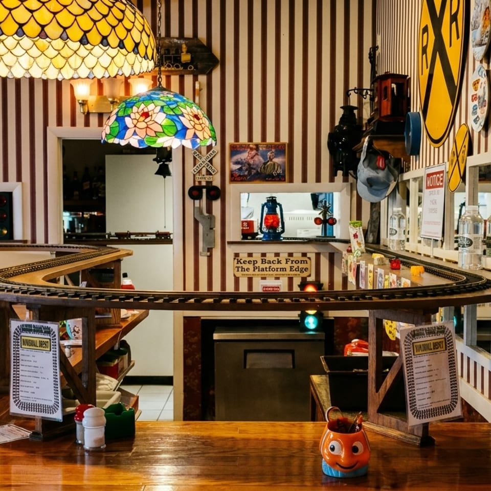 Restaurant interior with train tracks on a counter, striped walls, and stained-glass lamps.