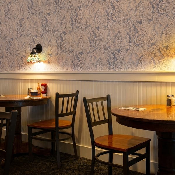 Cozy restaurant interior with round wooden tables, chairs, and stained-glass wall sconces illuminating patterned wallpaper.