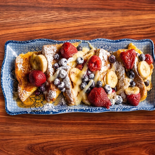 French toast with strawberries, blueberries, bananas, and powdered sugar on a blue and white plate.