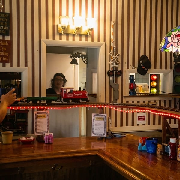 A man in a red shirt stands at a bar with a model train running on tracks above it.