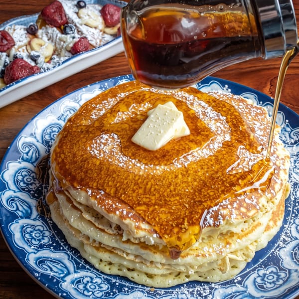 Maple syrup pouring over a stack of pancakes with butter, powdered sugar, and fruit on a blue plate.