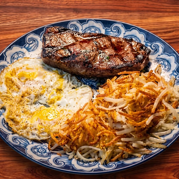 Grilled steak, two fried eggs, and crispy hash browns on a blue and white patterned plate.