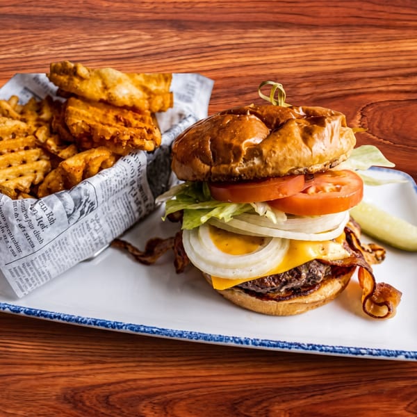 A cheeseburger with lettuce, tomato, onion, and bacon, served with waffle fries and a pickle.